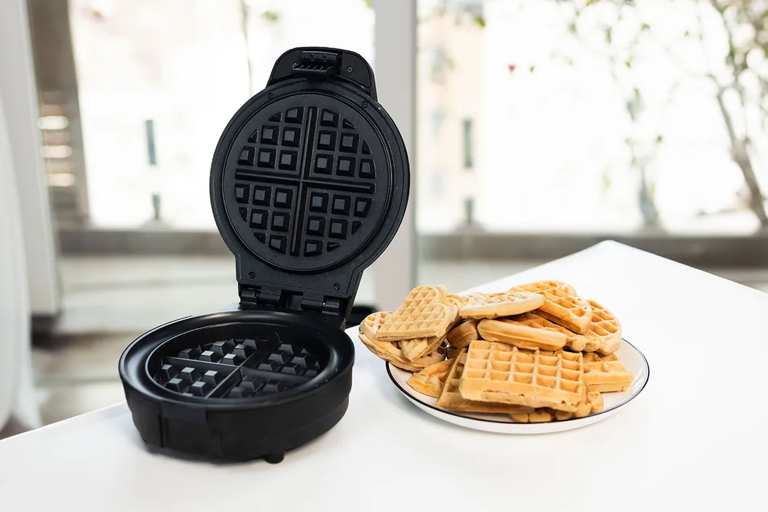 Chefman Anti-Overflow waffle maker with empty waffle plates on a white tabletop next to a plate containing stacks of waffles.