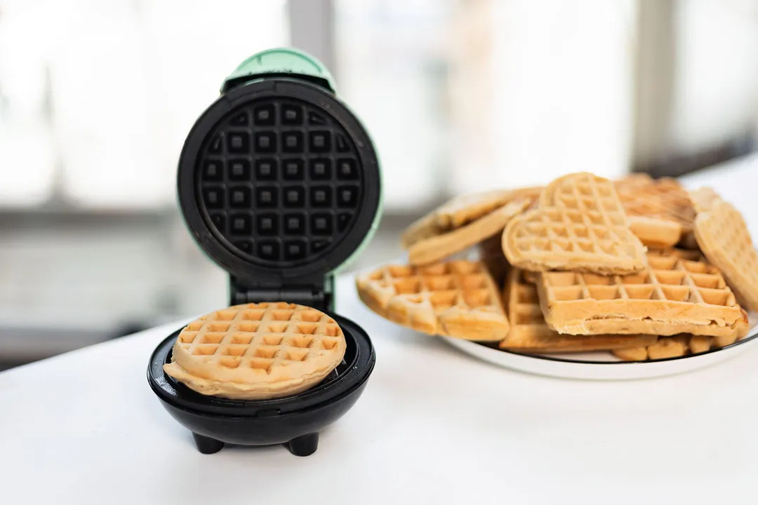 DASH Mini waffle maker after finishing cooking a mini-style waffle on a white countertop next to a plate of fresh, various-sized waffles.