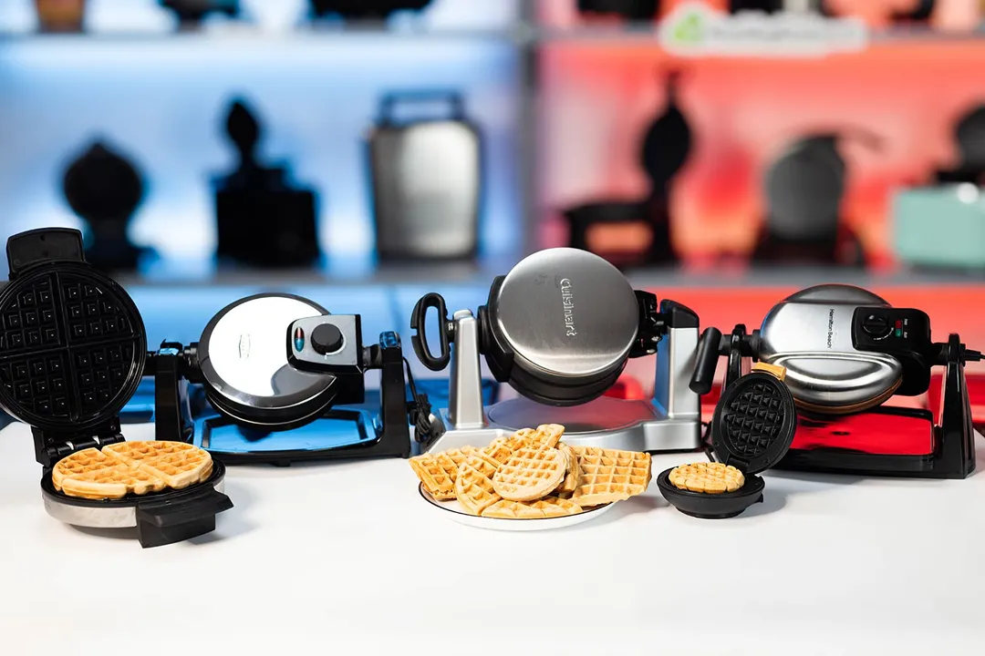 The best waffle makers on a white tabletop with plates of cooked waffles in front, set against a blurry red and blue background.