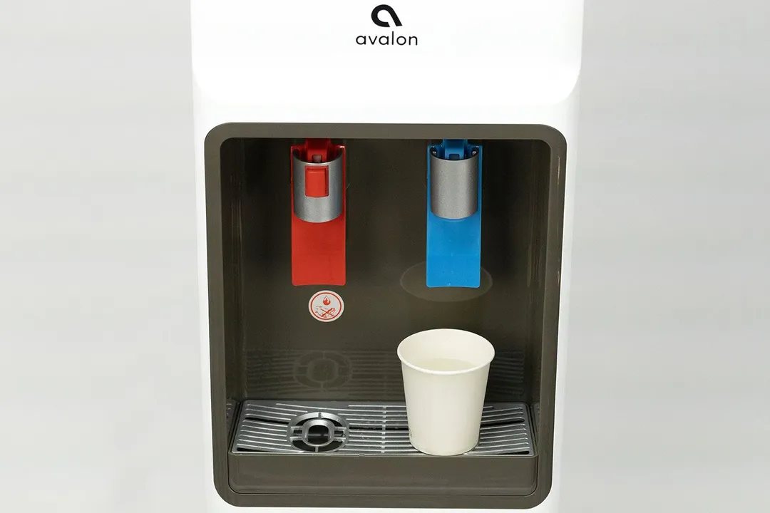 A paper cup filled with water resting on a drip tray below the cold water outlet on a water cooler dispenser.