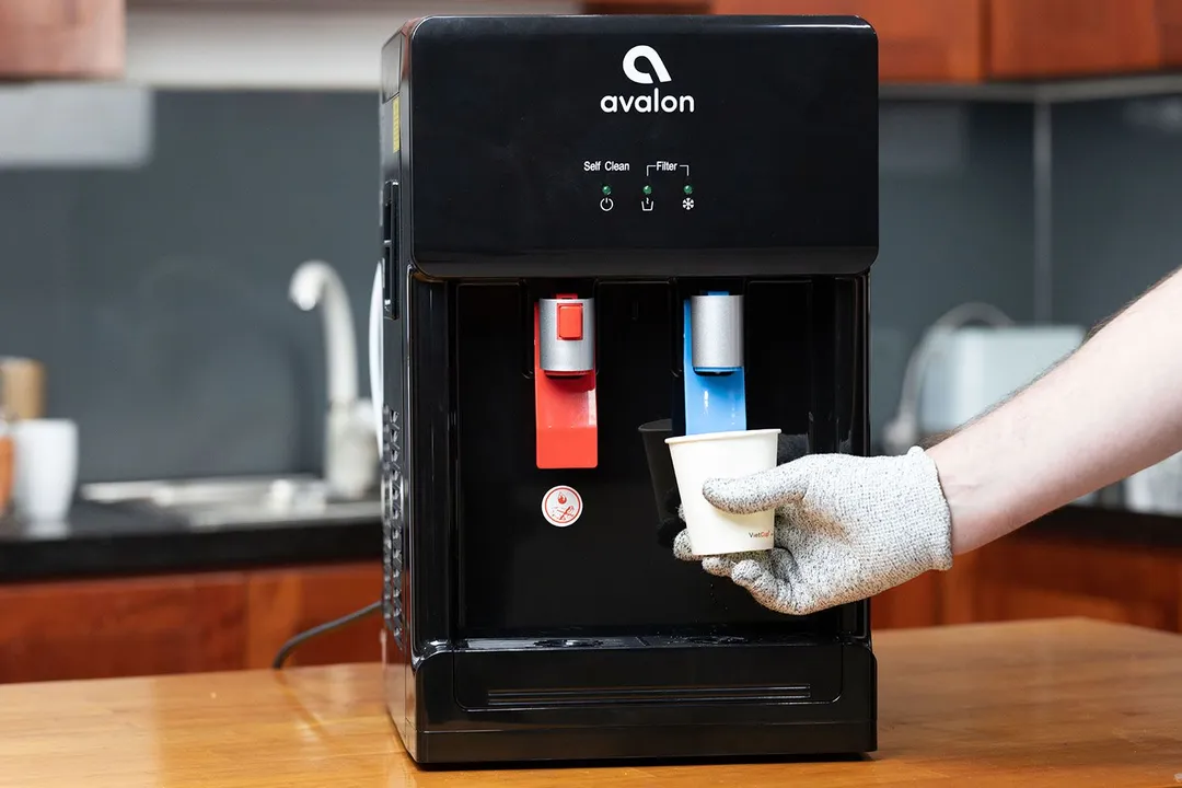 A gloved hand dispensing cold water from a water cooler dispenser into a part cup by pushing a blue lever.