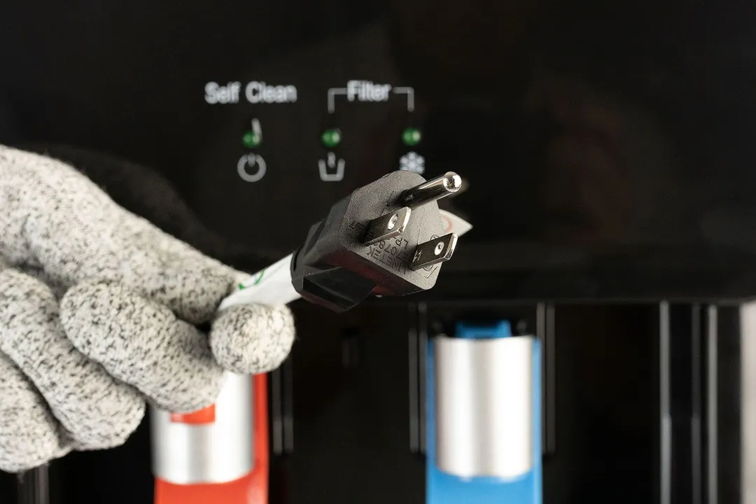 A gloved hand holding the three pronged plug of the Avalon A8 water cooler dispenser pictured against the control panel.