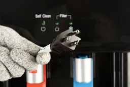 A gloved hand holding the three pronged plug of the Avalon A8 water cooler dispenser pictured against the control panel.