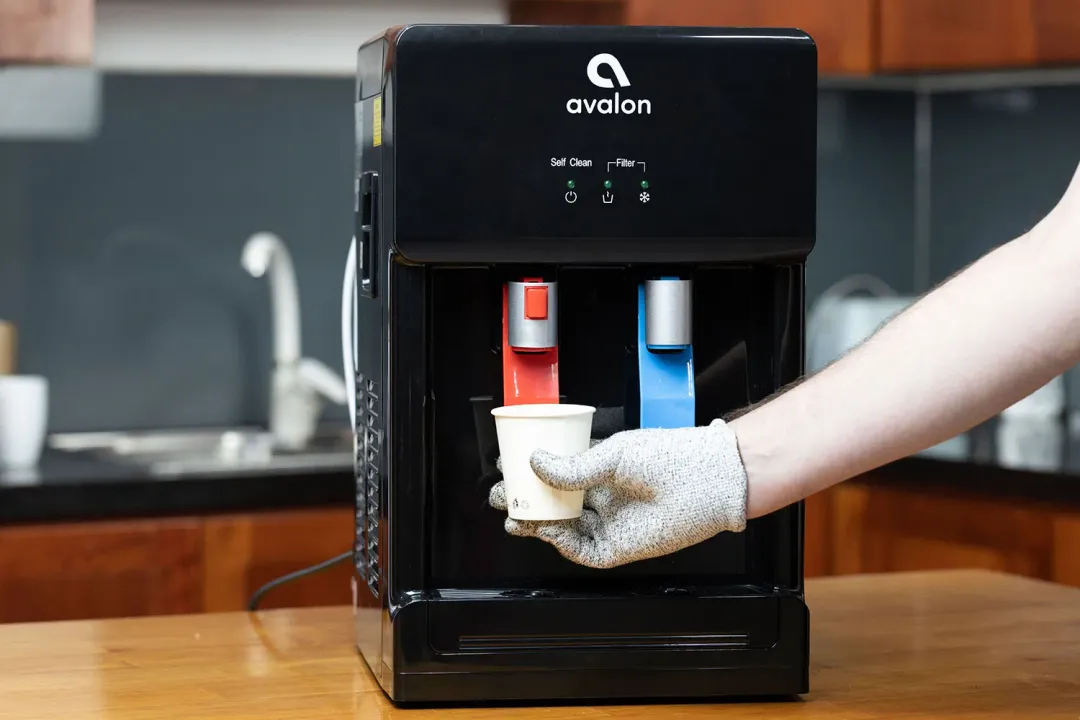  A gloved hand dispensing hot water from a water cooler dispenser into a paper cup.