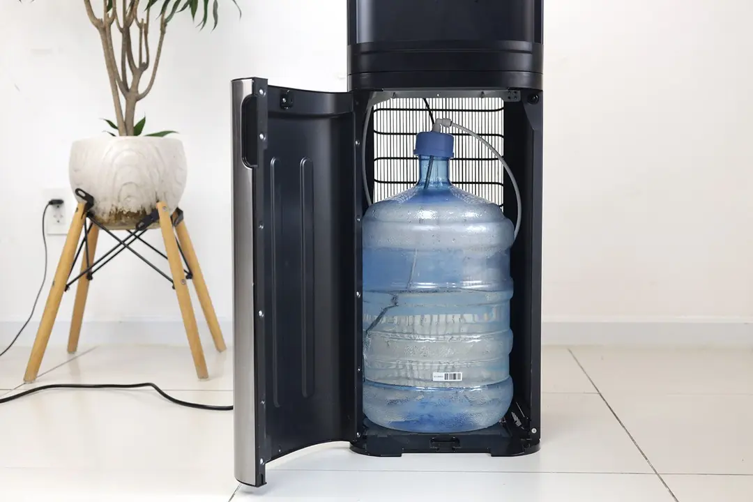 View of an open cabinet door of a bottom-loading water cooler dispenser with a full water bottle inside.