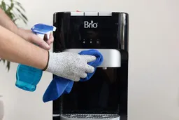 A hand holding a spray-bottle with cleaning liquid and another a cloth wiping the front panel of a water cooler dispenser.