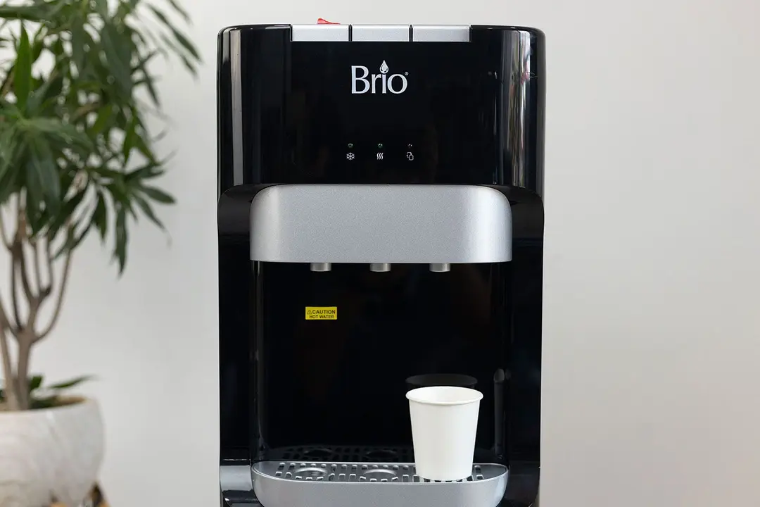A paper cup standing on the drip tray of a water cooler dispenser below the cold water spout.