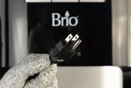 A gloved hand holding the three pronged plug of the Brio 400 water cooler dispenser up against the front control panel.