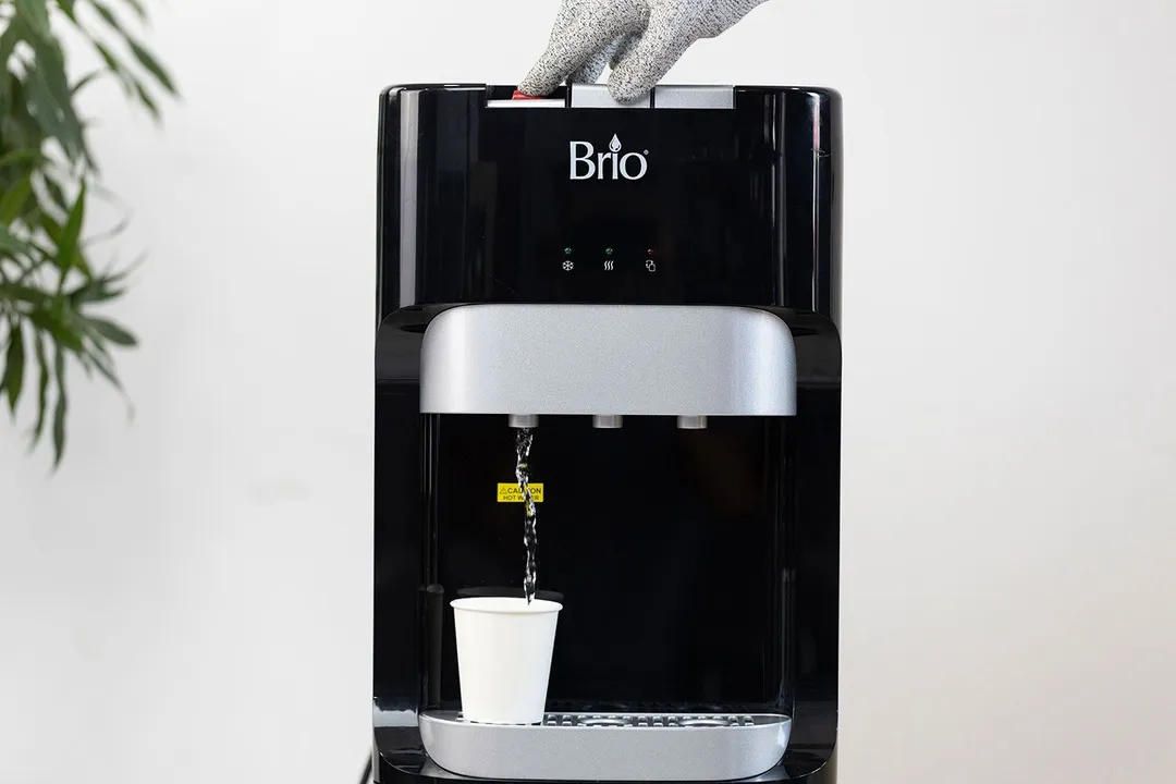 A paper cup resting on the drip tray of a water cooler dispenser while hot water is being dispensed.
