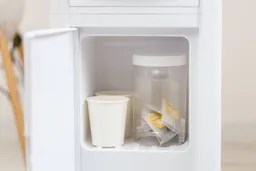 A small storage cabinet on a water cooler dispenser with two paper cups and plastic jar demonstrating the size.