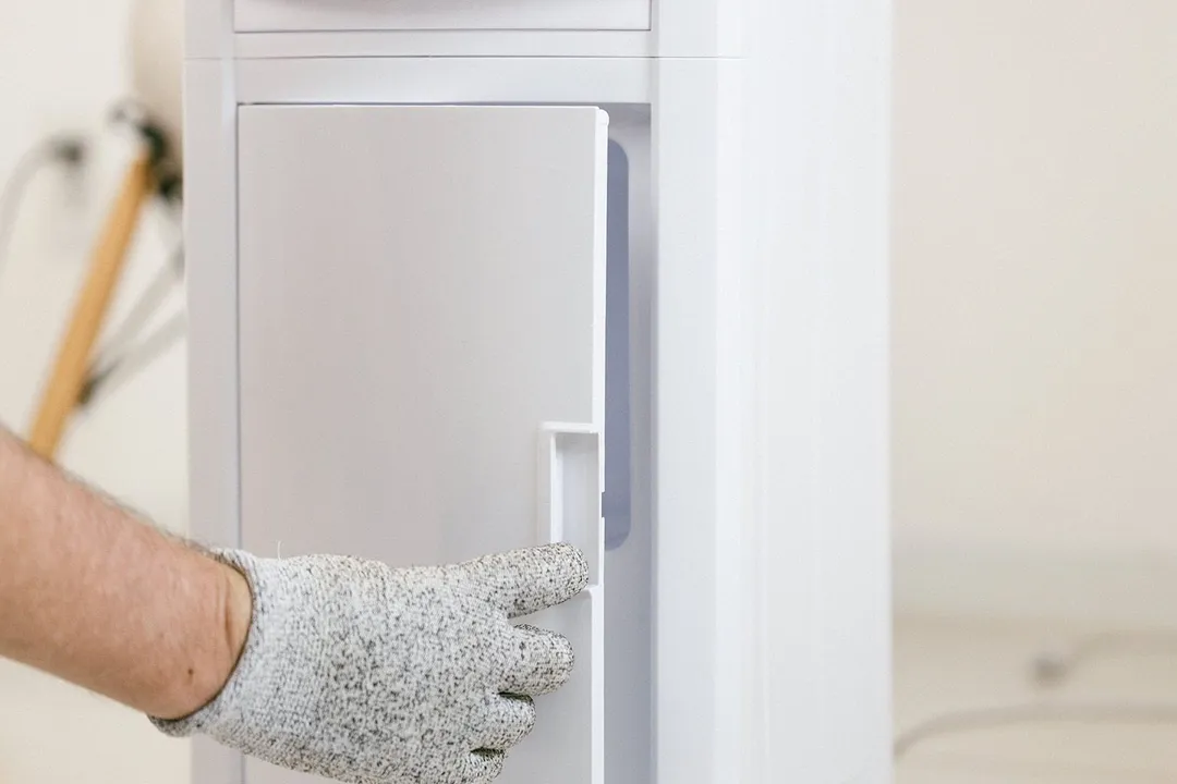 A gloved hand opening a small storage cabinet door of a top-loading water cooler dispenser.