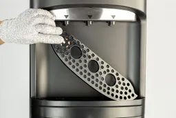 A gloved hand holding the lid of a trip tray across the front of a water cooler dispenser.