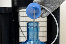A gloved hand holding a partly removed silicone bottle cap from a 5-gallon jug of a bottom-loading water cooler dispenser.