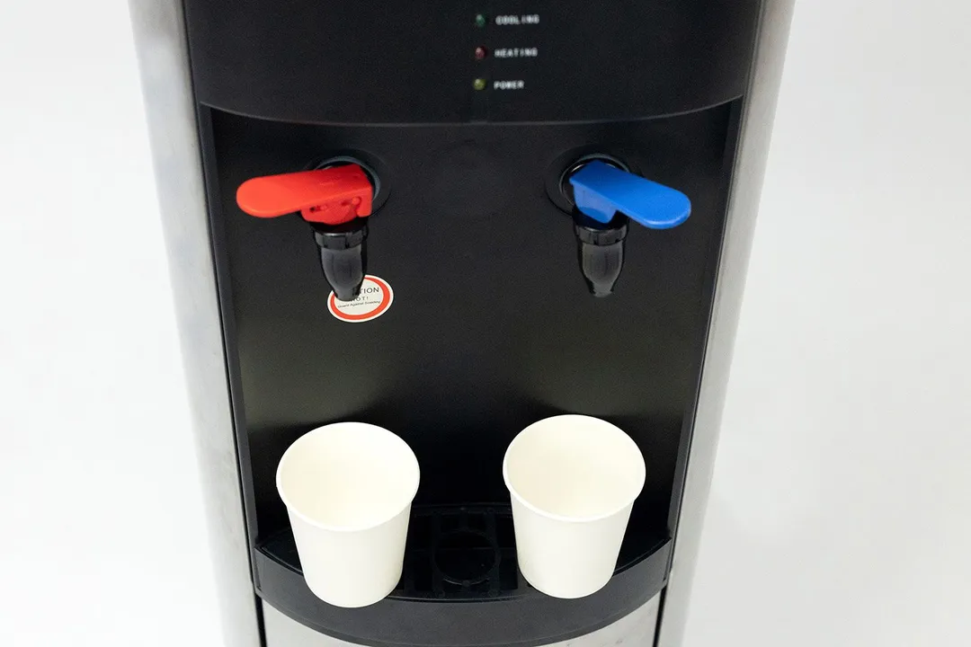 Two small paper cups barely fitting completely on the drip tray of a water cooler dispenser with a faucet design.