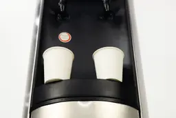 A bottom up view of two paper cups resting on the narrow drip tray of a water cooler dispenser.