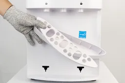 A gloved hand holding a drip tray of a water cooler dispenser with the cover askew and removed from its holding.