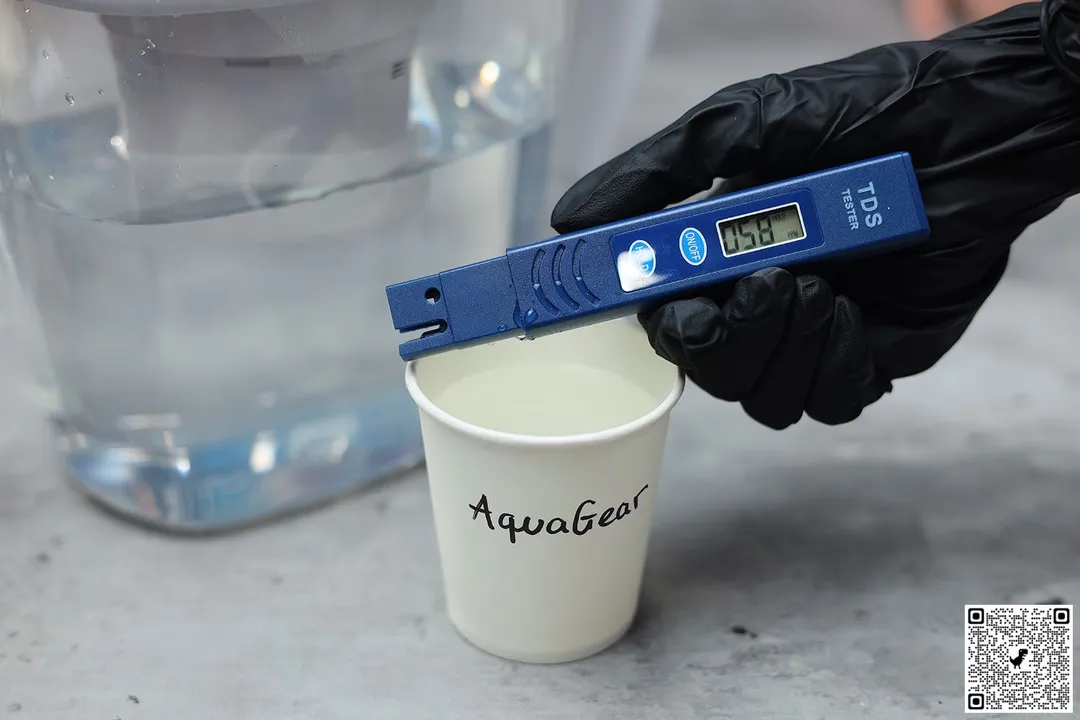 gloved hand holding water TDS meter over a paper cup labeled Aquagear next to lower part of water filter pitcher