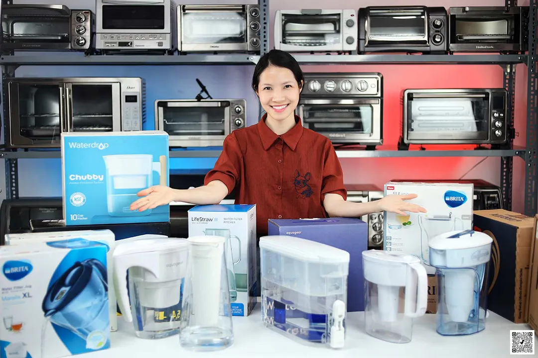 Woman spreading arms behind water filter pitchers and dispensers on a table, toaster ovens on a shelf at the back