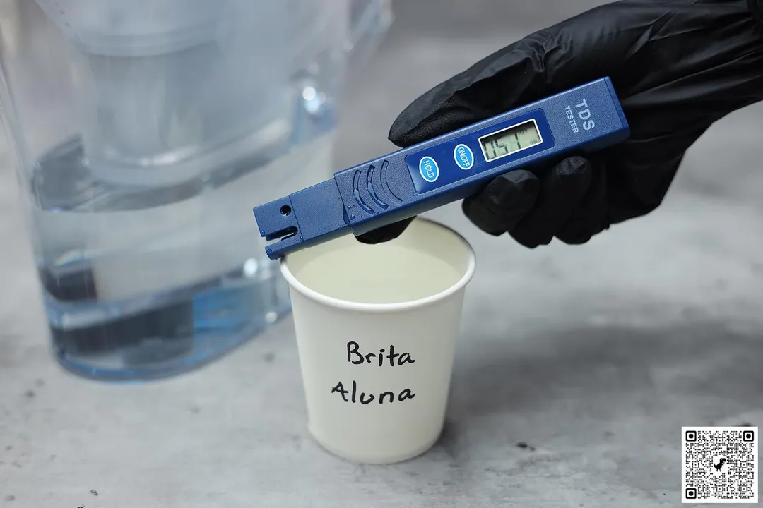 A gloved hand holding a water TDS meter over a paper cup with the words Brita Aluna written on it and the lower part of a water filter pitcher in the background