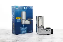 The silver, metallic Brita Basic faucet-mount water filtration system next to its shipping box.