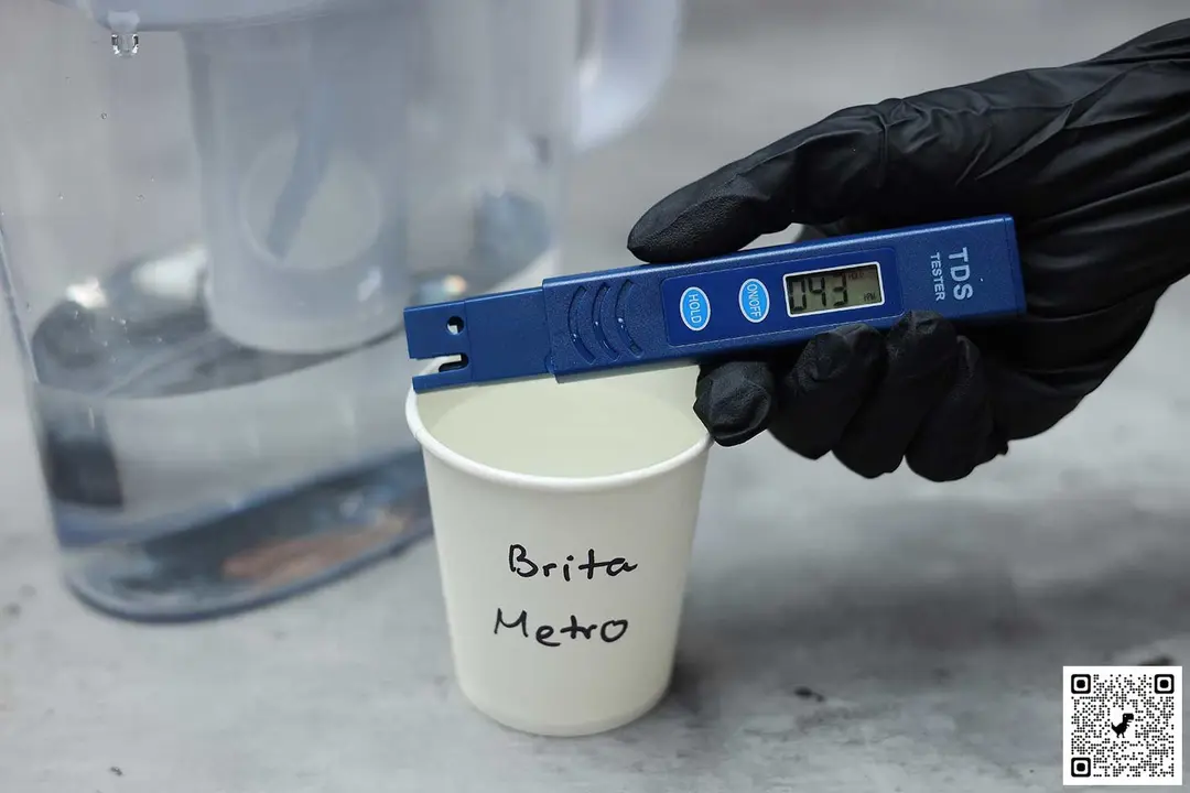Gloved hand holding a water TDS meter over a paper cup labeled Brita Metro, the lower part of a water filter pitcher in the background
