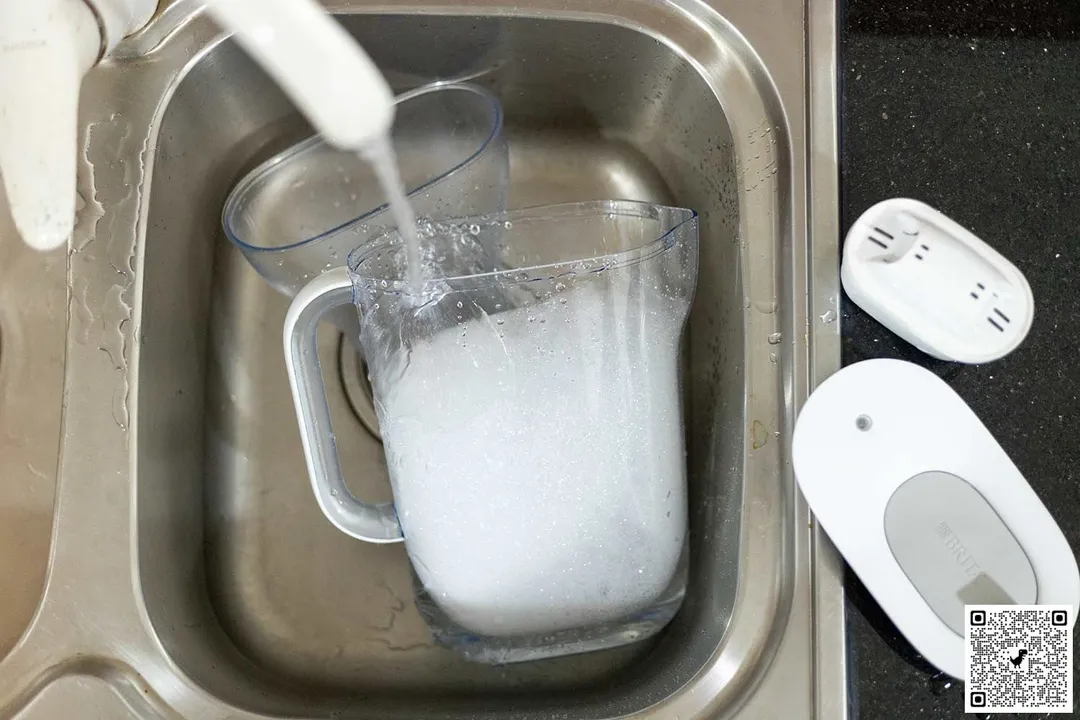 The Brita Style jug filled with soapy water and its reservoir in a sink, water running down from the faucet, the filter piece and jug lid on the left side of the sink