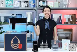 Woman standing, her hand pointing to the left where the Brita UltraMax XL sits on its package box, with smartphone, test tubes on a rack, and a large glass of water on the table in front of her