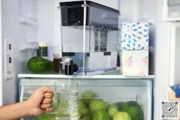 Hand holding a glass under the running spigot of the Brita UltraMax XL 27 cup dispenser, which is placed in a fridge along with fruits and paper packages