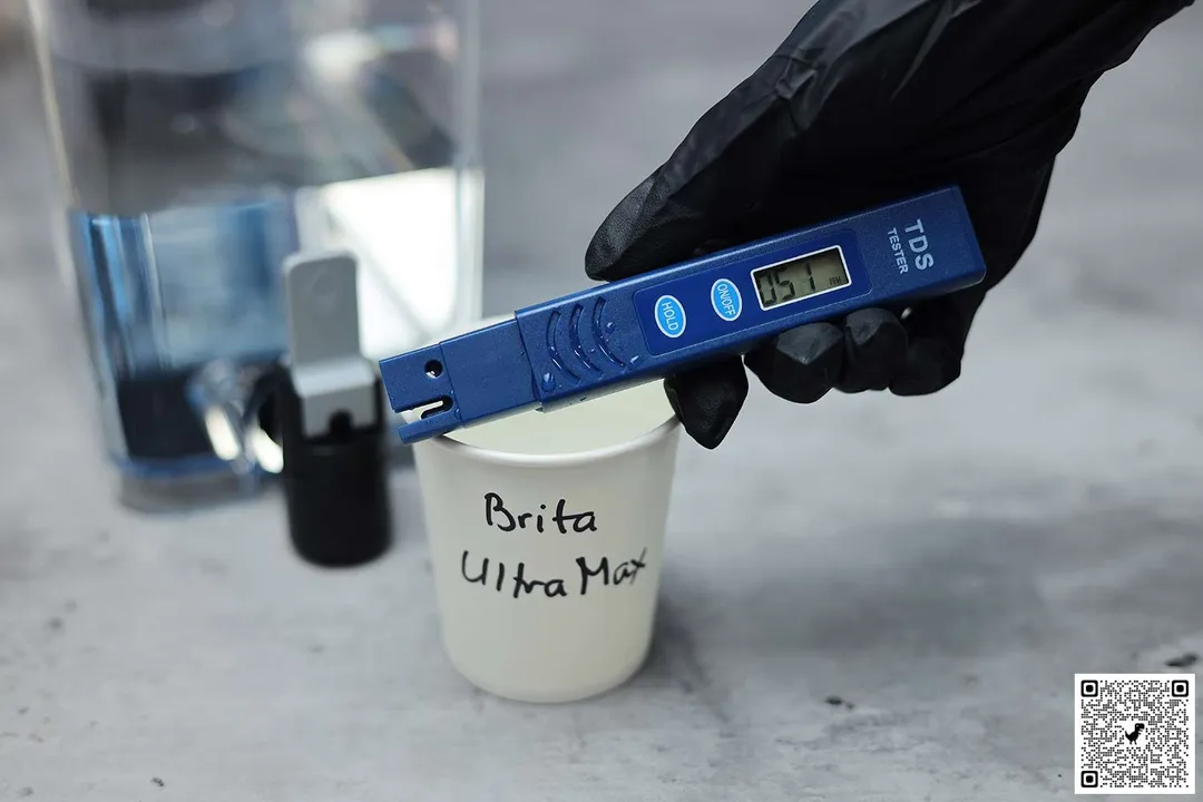 Gloved hand holding a water TDS meter over a paper cup labeled Brita UltraMax, next to the lower part of the water dispenser