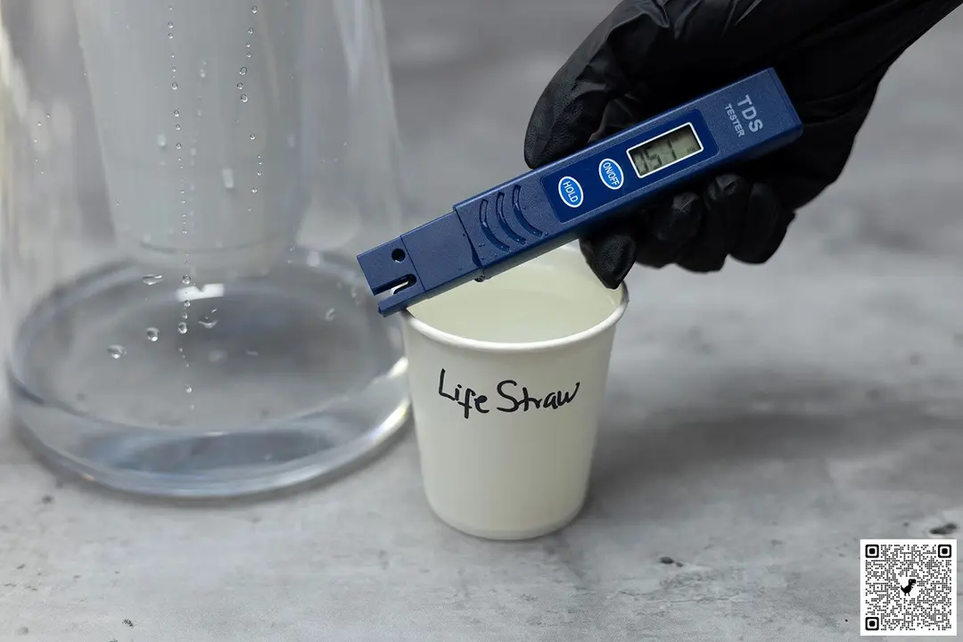 Gloved hand holding a water TDS meter over a paper cup labeled LifeStraw, the lower part of a water filter pitcher in the background
