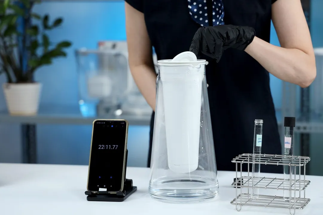 Person checking a water filter pitcher on a table along with test tubes on a rack and a smartphone timer