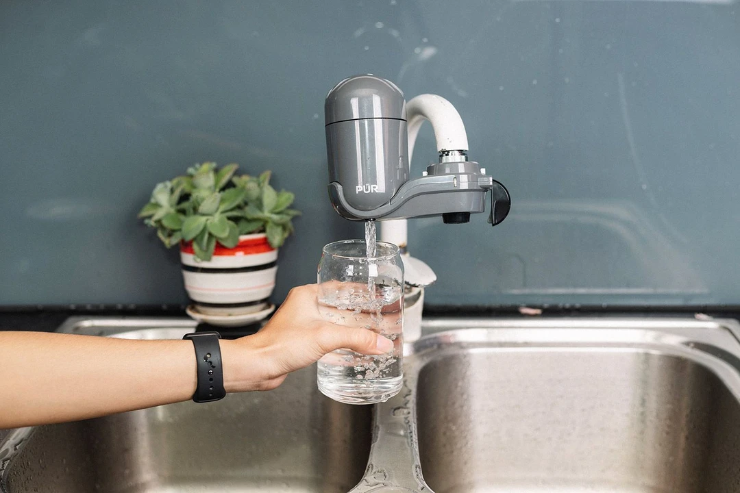 The grey, plastic PUR FM2500V faucet-mount filter mounted on a kitchen faucet dispensing clean water into a cup underneath.