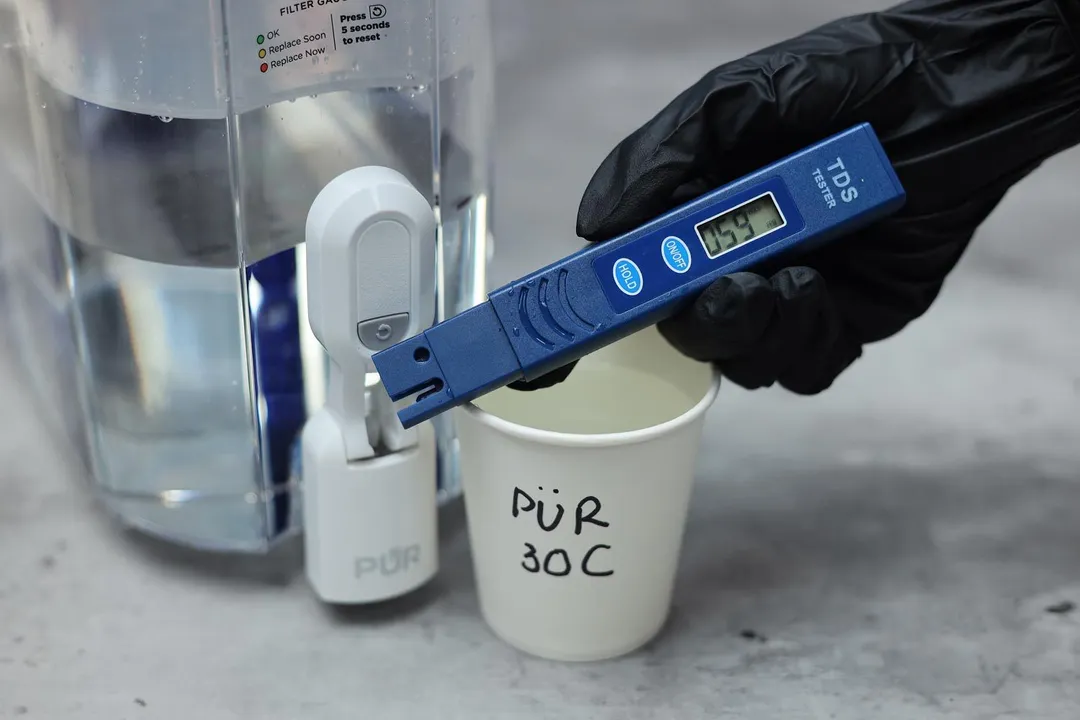 Gloved hand holding a water TDS meter over a paper cup labeled PUR 30C, next to the water dispenser