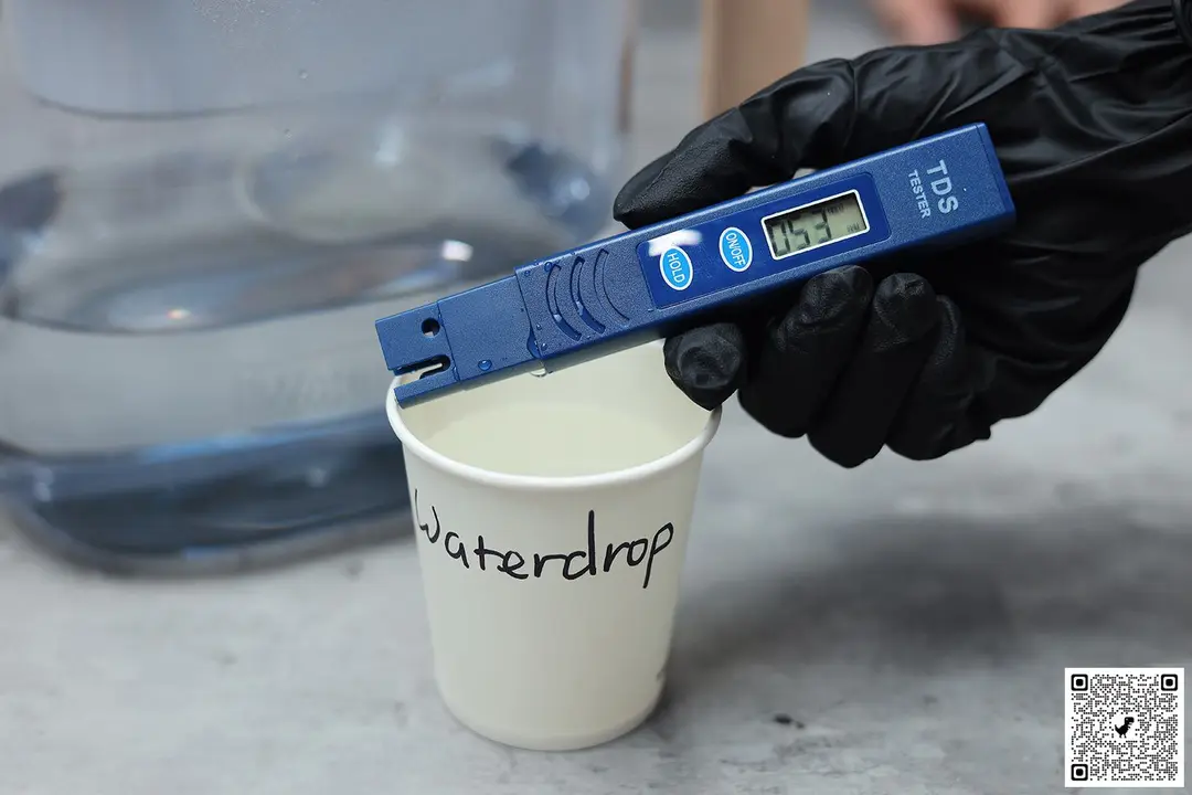 Gloved hand holding TDS meter over a paper cup labeled Brita Aluna and the lower part of a water filter pitcher