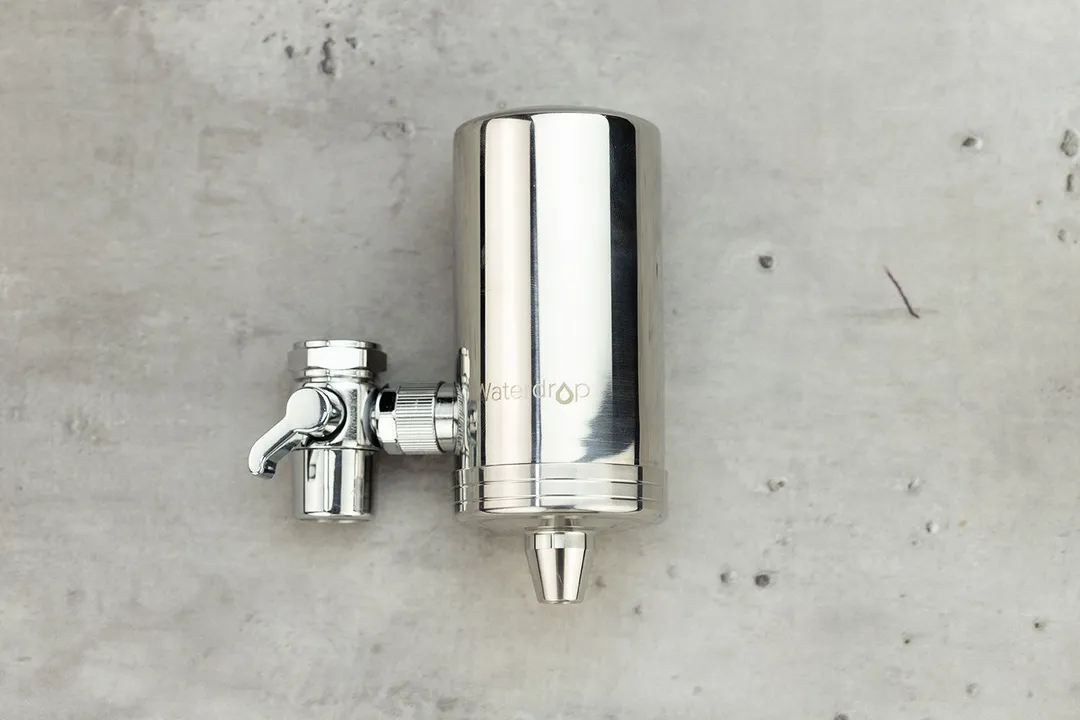 The silvery, metallic Waterdrop WD-FC-06 faucet-mount water filter on a grey table.