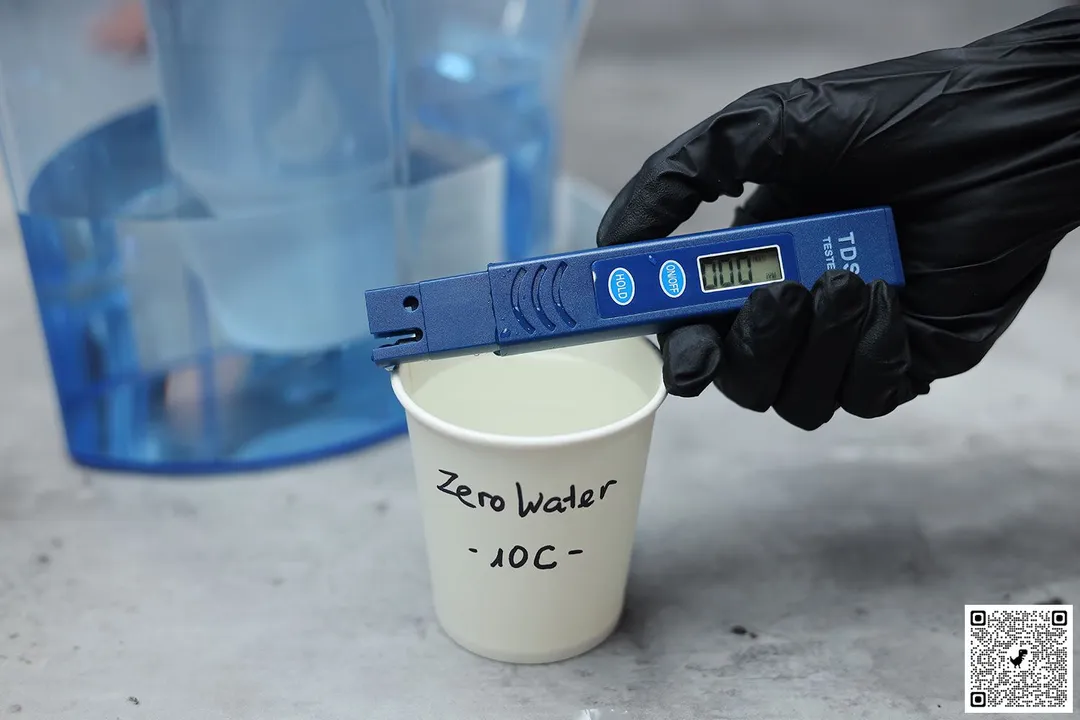 Gloved hand holding TDS meter over a paper cup labeled ZeroWater 10C and the lower part of a water filter pitcher